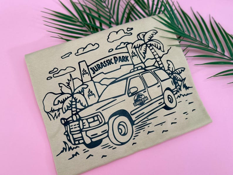 May include: A brown t-shirt with a black line drawing of a Jeep driving through a jungle with palm trees and the words "Jurassic Park" on a sign.