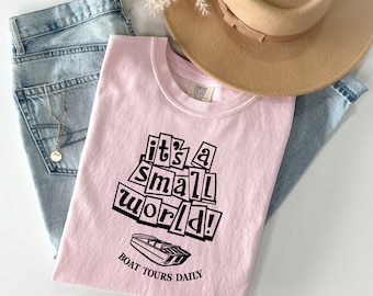 Może przedstawiać: Różowy T-shirt z czarnym nadrukiem, na którym widnieje napis "it's a small world!" oraz czarno-biała ilustracja łodzi z napisem "Boat Tours Daily" pod spodem.