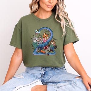 Puede incluir: Camiseta verde oliva con un gr&aacute;fico de dibujos animados de personajes de animales, incluyendo un zorro, un conejo y un castor, con el n&uacute;mero "2" en un dise&ntilde;o azul en forma de serpiente. Los nombres de los personajes est&aacute;n escritos en la camiseta.