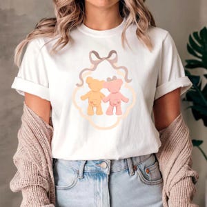Könnte beinhalten: Weißes T-Shirt mit einer Grafik von zwei Teddybären, einem gelben und einem rosa, die sich in einem dekorativen Rahmen an den Händen halten. Über den Bären befinden sich eine hellbraune Schleife und Bänder. Das T-Shirt hat kurze Ärmel und umgeschlagene Manschetten.