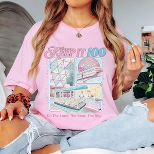 Op de afbeelding: Een lichtroze t-shirt met de tekst "Keep It 100" en illustraties van een pretpark. Het shirt heeft een retro-ontwerp met afbeeldingen van een geodetische bol, een monorail en een boot. De tekst "On The Land, The Seas, The Sky" staat eronder.