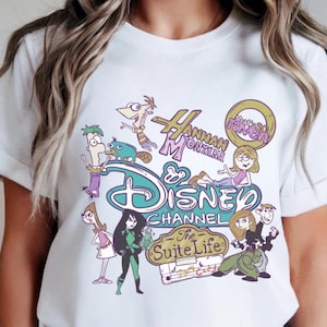 May include: White t-shirt featuring a colorful graphic design with cartoon characters and logos from Disney Channel shows, including "Hannah Montana" and "The Suite Life". The design includes the Disney Channel logo.