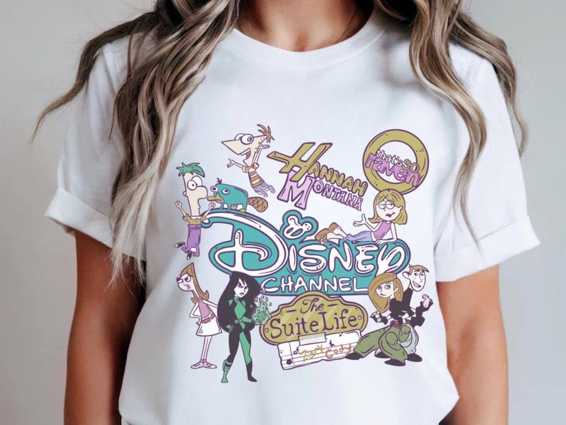 May include: White t-shirt featuring a colorful graphic design with cartoon characters and logos from Disney Channel shows, including "Hannah Montana" and "The Suite Life". The design includes the Disney Channel logo.