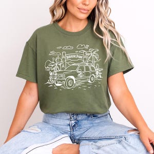 May include: A green t-shirt with a white graphic of a jeep driving through a jungle with the text "Jurassic Park" on a sign.