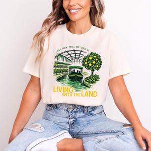 以下が含まれることがあります： 「Living with the Land」の文字が入った、緑と黄色のグラフィックが特徴のクリーム色のTシャツ。デザインには、水路を走るボート、温室、果樹が含まれています。このシャツは半袖で、クルーネックです。