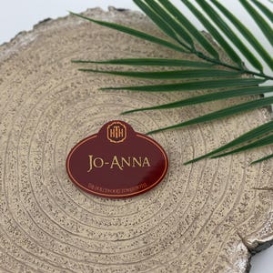 Op de afbeelding: Een bordeauxrode naamplaat met gouden letters en een gouden embleem. De naamplaat leest "Jo-Anna" en "The Hollywood Tower Hotel". De tag is ovaal van vorm en rust op een lichtbruin, getextureerd oppervlak met een groen blad.
