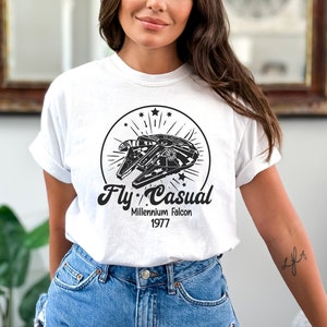 Può includere: Maglietta bianca con una grafica in bianco e nero del Millennium Falcon con il testo "Fly Casual" e "Millennium Falcon 1977" sotto.