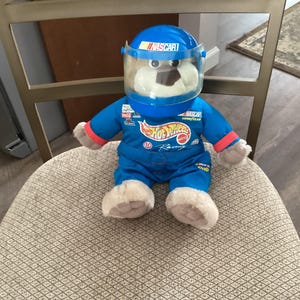 May include: A blue and white plush toy wearing a blue helmet with the word "NASCAR" on it. The toy is wearing a blue racing suit with the Hot Wheels logo and the words "Hot Wheels Racing".