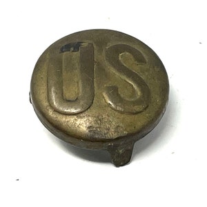 May include: A brass button with the letters "US" embossed on the front. The button has a raised edge and a flat back.