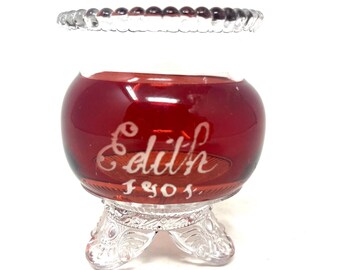 May include: A red glass bowl with a clear glass base and three legs. The bowl has a white inscription that reads "Edith 1904."