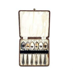 May include: A set of six silver teaspoons in a brown leather case. The spoons are arranged in a row and are all facing the same direction. The case is open and the spoons are visible.