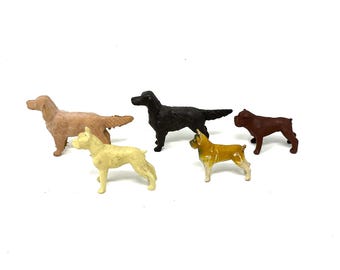 May include: Five small dog figurines in various colors, including tan, black, yellow, and brown. The dogs are in different poses, with some standing and others in a walking position. The figurines are on a white background.
