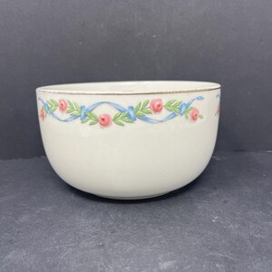 May include: A white ceramic bowl with a gold rim and a blue and pink floral design. The design features pink roses and blue ribbons.