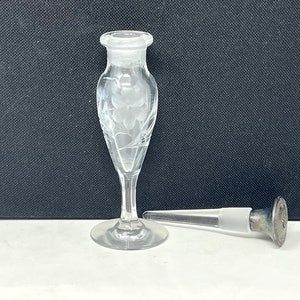 May include: Clear glass perfume bottle with etched floral design. The bottle has a long, slender stem and a round base. A separate stopper with a silver-colored metal top is next to the bottle.