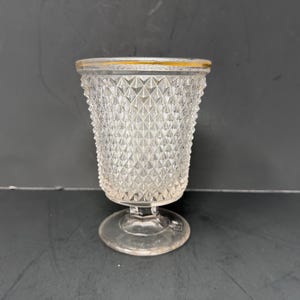 May include: Clear glass goblet with a diamond pattern and a gold rim. The goblet has a pedestal base and a flared top. The glass is transparent, allowing light to pass through. The goblet is likely vintage or antique.
