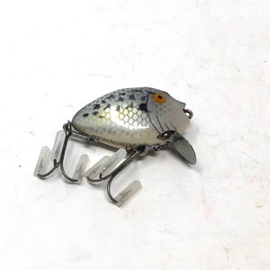 May include: A silver and black fishing lure with a white and black fish design. The lure has a single hook and the text "Plunger" is printed on the side.