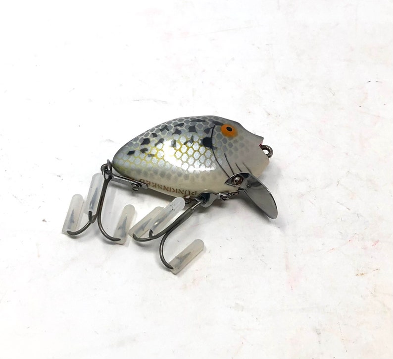 May include: A silver and black fishing lure with a white and black fish design. The lure has a single hook and the text "Plunger" is printed on the side.