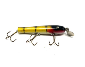 Pode incluir: Um isco de pesca de madeira amarelo, preto e vermelho com três anzóis triplos. O isco tem um design de peixe realista com um olho preto e uma cauda prateada.