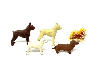 May include: A collection of small dog figurines in various breeds and colors. The figurines include a brown Boxer, a cream-colored Boxer, a white Bulldog, a brown Dachshund, and a multi-colored fluffy dog.