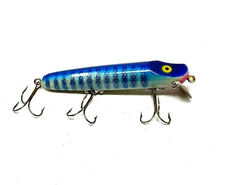 Può includere: Un'esca da pesca blu e bianca con un occhio giallo e labbra rosse. L'esca ha una finitura argentata e tre ami tripli.
