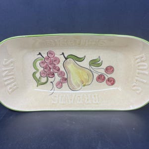 May include: A white ceramic serving dish with a green rim. The dish has a pear, grapes, and cherries painted on it. The words "BREADS", "ROLLS", "BISCUITS", and "RUINS" are written on the dish.