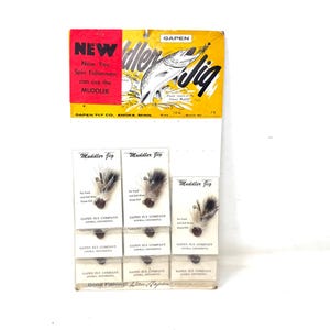 May include: A vintage Gapen Muddler Jig retail display card featuring a yellow and white background with a fish illustration and text that reads NEW Now You Spin Fishermen can use the MUDDLER. The card holds several fishing lures with brown heads.