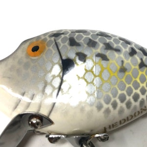 May include: A white and gray fishing lure with a black and yellow eye. The lure has a silver finish and a black and gray pattern. The lure is made by Heddon.