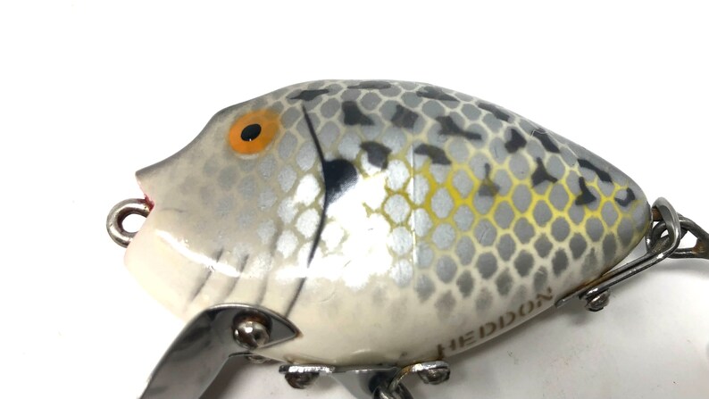 May include: A white and gray fishing lure with a black and yellow eye. The lure has a silver finish and a black and gray pattern. The lure is made by Heddon.