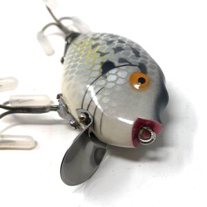 May include: A white and gray fishing lure with a black and orange eye. The lure has a silver propeller and three treble hooks.