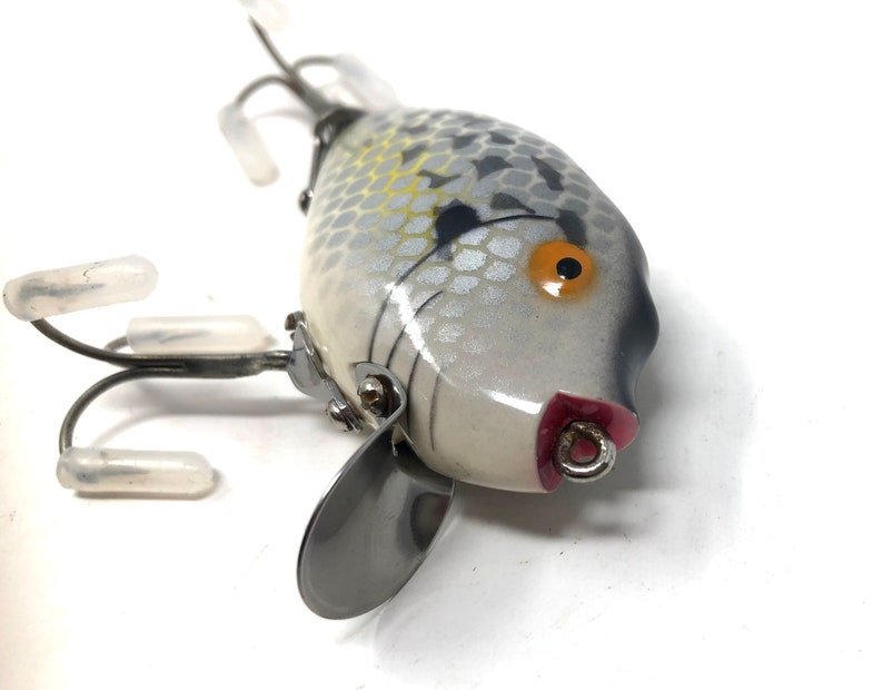 May include: A white and gray fishing lure with a black and orange eye. The lure has a silver propeller and three treble hooks.