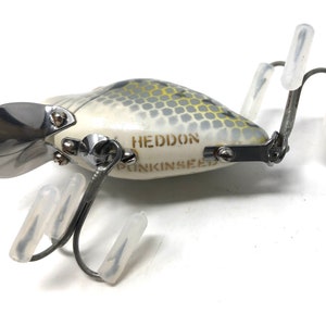 May include: A white and grey fishing lure with a silver propeller and the text "HEDDON PUNKINSEED" printed on the side. The lure has three hooks with clear plastic covers.