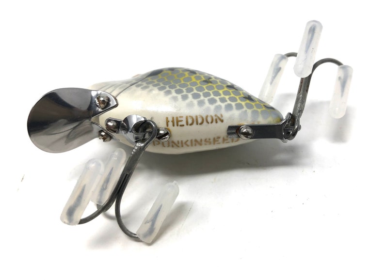 May include: A white and grey fishing lure with a silver propeller and the text "HEDDON PUNKINSEED" printed on the side. The lure has three hooks with clear plastic covers.
