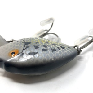 May include: A silver and black fishing lure with a realistic fish design. The lure has a yellow and orange eye and a textured body.
