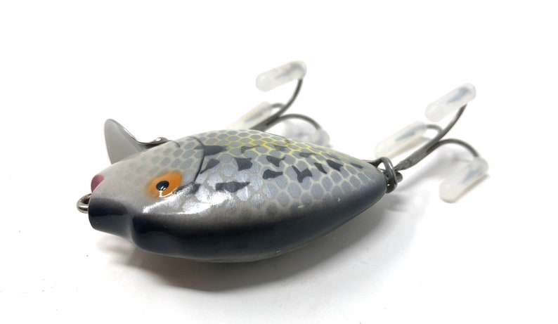 May include: A silver and black fishing lure with a realistic fish design. The lure has a yellow and orange eye and a textured body.
