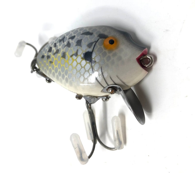 May include: A white and gray fishing lure with a yellow eye and red mouth. The lure has a silver finish and a textured surface. It has three treble hooks and a swivel.