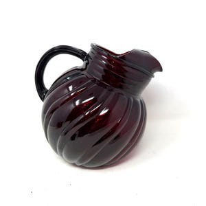 May include: A vintage, ruby-red glass pitcher with a black handle. The pitcher has a spiral design and a spout for pouring. The handle is a curved, solid piece. The pitcher is likely used for serving beverages.