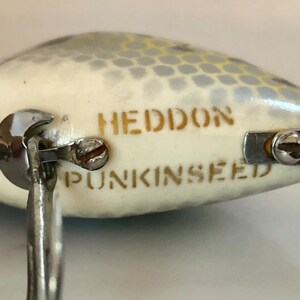 May include: A white fishing lure with a silver and yellow pattern. The lure has the words "HEDDON" and "PUNKINSEED" printed on it.