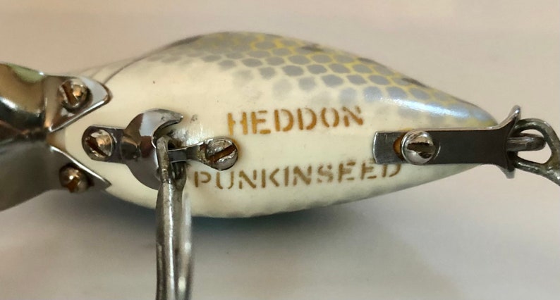 May include: A white fishing lure with a silver and yellow pattern. The lure has the words "HEDDON" and "PUNKINSEED" printed on it.