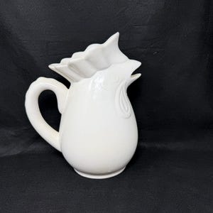 May include: A white ceramic pitcher shaped like a rooster. The vessel features a curved handle on the side, a molded comb on the top, and a beak-shaped spout. The surface is smooth and glossy, with embossed details defining the bird's facial features.