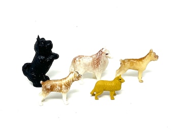 May include: A collection of five miniature dog figurines in various breeds and poses. The dogs are painted in shades of black, tan, and yellow, with white accents. The figurines are standing on a white surface.