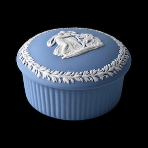 Puede incluir: Caja de baratijas de cerámica azul con un diseño en relieve blanco en la tapa. La tapa presenta una escena clásica con figuras, rodeada por una guirnalda de hojas blancas. La caja tiene una base acanalada y un acabado liso y mate.