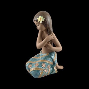 Puede incluir: Una figura de porcelana de una persona arrodillada con cabello castaño, que viste un pareo color verde azulado con rayas doradas y motivos florales. La figura tiene una flor amarilla pálida tras la oreja y toca su cabello con una mano.