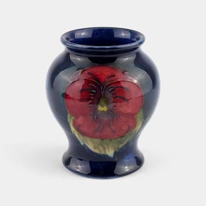 May include: A dark blue ceramic vase with a glossy finish. The vase features a large, detailed red pansy flower design with green leaves. The vase has a rounded body and a flared rim, suitable for displaying flowers.
