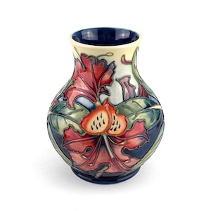 May include: A decorative ceramic vase with a dark blue interior and base. The vase is adorned with a vibrant floral pattern of red, green, and blue leaves, and a cream-colored rim. The design includes two orange seed pods.