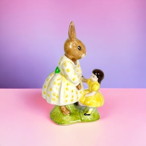 Puede incluir: Una figurita de cerámica de un conejo con un vestido amarillo con lunares blancos y un lazo verde. El conejo está sosteniendo la mano de una niña pequeña que lleva un vestido amarillo con lunares blancos. La figurita está sobre un fondo rosa y morado.