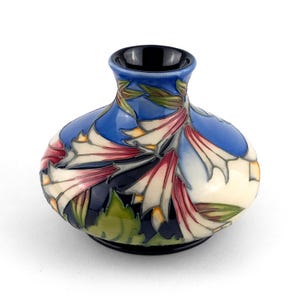 May include: A small, round ceramic vase with a black rim and base. The vase has a blue background with a floral design in white, pink, and green. The flowers are stylized and detailed, creating a decorative piece.