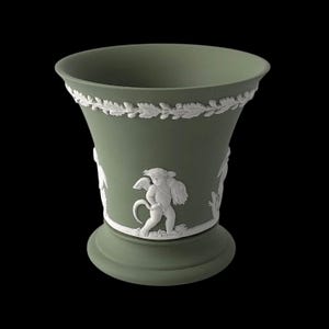 Op de afbeelding: Een saliegroene keramische vaas met witte reliëfdetails. De vaas heeft een decoratieve band van bladeren rond de rand en cherubijnfiguren. De vaas heeft een uitlopende bovenkant en een ronde basis, tegen een zwarte achtergrond.