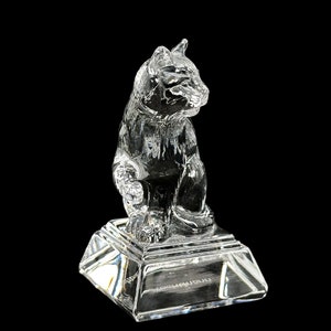May include: A clear crystal figurine of a cat sitting on a square base. The cat is detailed with a textured fur pattern and has a realistic pose.