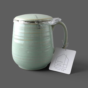 May include: A light green ceramic infuser mug with a matching lid, featuring a reactive glaze finish and horizontal ridges. The mug is wrapped in clear plastic and includes a white tag with the text SCANDI HOME and Reactive Glaze Infuser Mug.