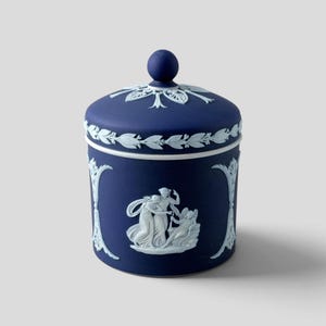 May include: A dark blue cylindrical jar with a matching lid featuring a spherical knob. The exterior is decorated with white relief carvings of classical figures and floral motifs, including a central scene of three figures and vertical accents.